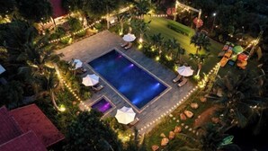 Un área de piscina con una pileta rectangular, rodeada de sillones de descanso y sombrillas, con un sendero bien iluminado y luces decorativas.