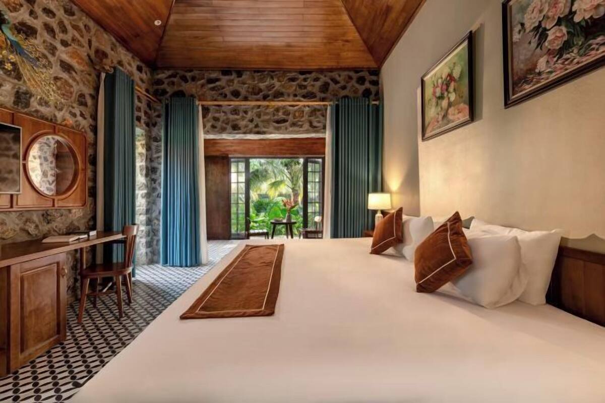 A bedroom with a stone wall, a wooden ceiling, a bed with pillows, a desk with a chair, and a view of greenery outside.