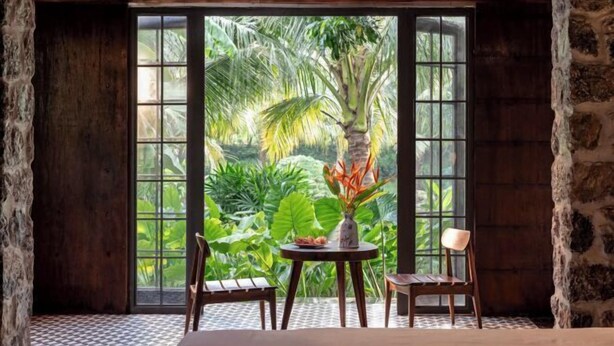 Una habitación con un ventanal grande, una mesa con sillas y vistas a plantas tropicales.
