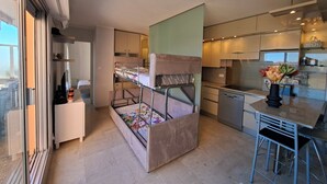 A modern kitchen with a built-in stove, a dining area with a bar stool, and a bunk bed with a colorful bedspread. - Quiet apt, A/C, 10mn walk to beaches, large terrace w/view over old town (Menton)