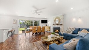 Una sala de estar moderna con un sofá azul, una mesa de centro de vidrio y una chimenea. Hay un comedor con una mesa y sillas, y una cocina con una encimera blanca y taburetes.