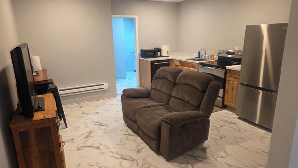 A modern living room with a reclining chair, a wooden entertainment unit, a flat-screen TV, and a kitchen area with a refrigerator, stove, and microwave. - Man cave 2.0 (Circle)