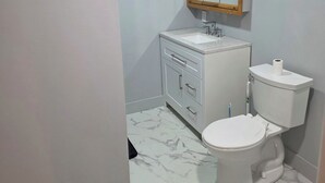 A bathroom with a white toilet, a white sink with a wooden frame mirror, and marble-patterned flooring. - Man cave 2.0 (Circle)