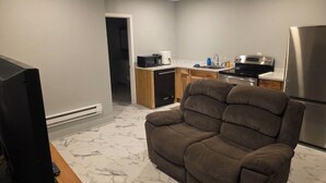 A living room with a brown sofa, a TV, and a kitchen area with a microwave, stove, and refrigerator. - Man cave 2.0 (Circle)
