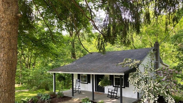 A small, single-story house with a porch, surrounded by lush greenery and trees.