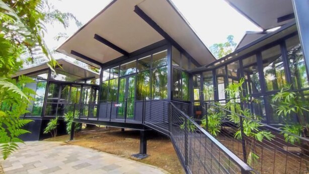 Un bâtiment moderne doté de grandes fenêtres en verre, d’une allée couverte et entouré de verdure.