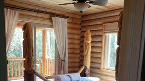 Une chambre située dans une cabane en rondins, avec un lit en bois, un ventilateur de plafond et de grandes fenêtres.