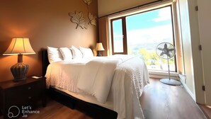 Une chambre avec un grand lit, deux lampes de chevet, un ventilateur fixé au mur et une fenêtre donnant sur des arbres.