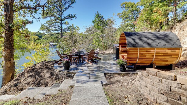 A wooden boat-shaped structure with a built-in grill, surrounded by a patio with a table and chairs, set against a backdrop of trees and a body of water. - Grand Lake Waterfront Getaway w/ Boat Slip! (Eucha)