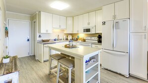 A compact kitchen with white cabinets, a central island with stools, a microwave, and a refrigerator. - Steps to Sea! 3rd-Floor Myrtle Beach Condo w/ Pool (Myrtle Beach)