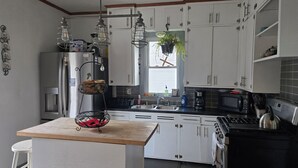 Una cocina con gabinetes blancos, un refrigerador de acero inoxidable, un microondas y una estufa. Hay una isla de madera con una cesta y una planta colgante.