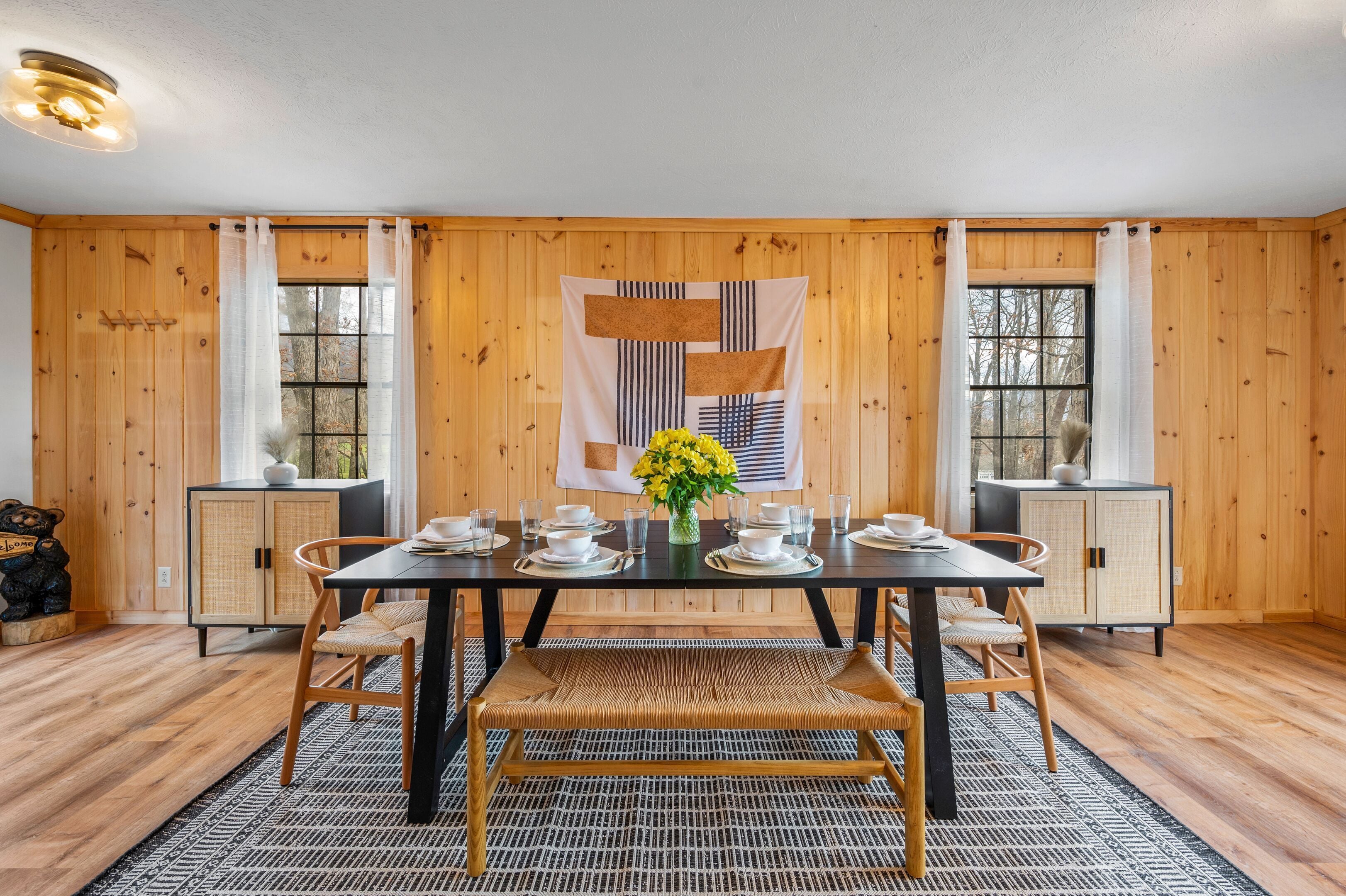Un coin repas avec une table en bois dressée pour quatre personnes, entourée d’armoires en bois, d’un tapis à motifs et d’une tapisserie murale.