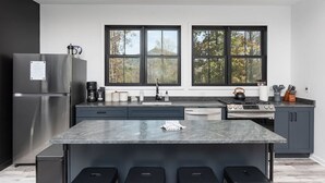 Une cuisine moderne dotée d’un îlot central, de meubles de rangement aux couleurs sombres et d’une grande fenêtre offrant une vue sur les arbres.