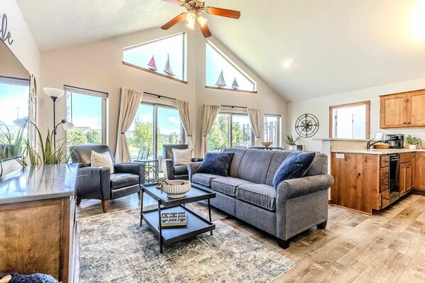 A spacious living area with a grey sofa, a coffee table, and a dining table. There are large windows with curtains, a ceiling fan, and a kitchen area visible.
