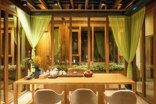 A dining area with a wooden table, chairs, and a view of greenery through large windows. - Anju·Rizhao Jinshan Designers' Panoramic Rooftop Cafe and Snow Mountain View (Lijiang)