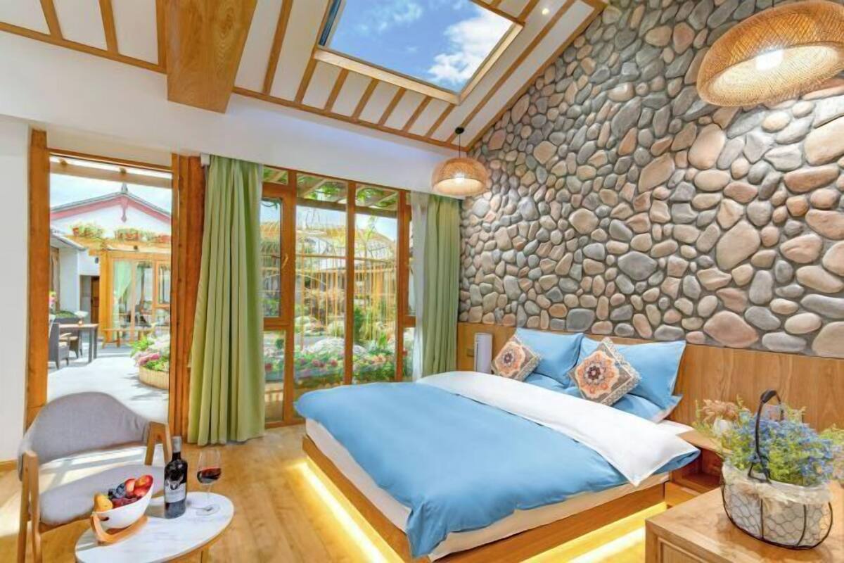 A bedroom with a stone wall, a bed with blue bedding, a dining table with fruit and wine, and a view of the outside.