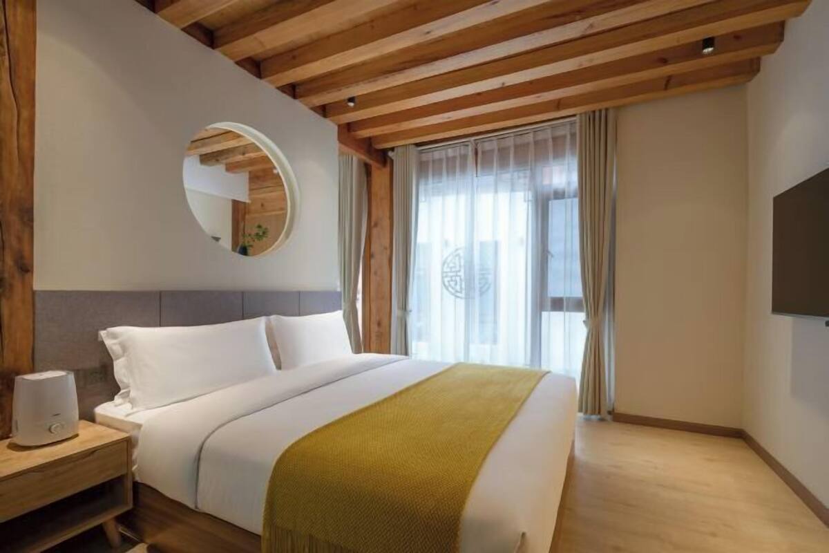 A hotel room with a wooden ceiling, a bed with white bedding, a bedside table with a lamp, and a TV mounted on the wall.