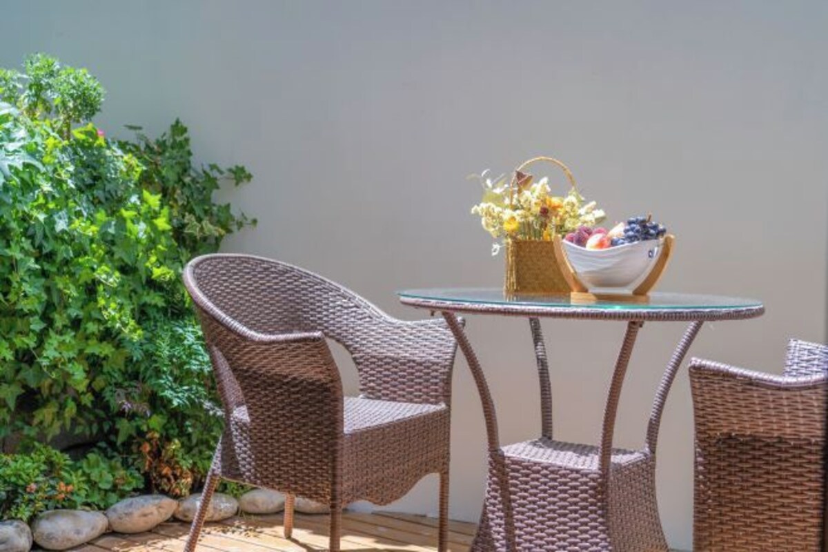 A wicker outdoor seating area with a glass table and chairs, a fruit bowl, and a basket of flowers.