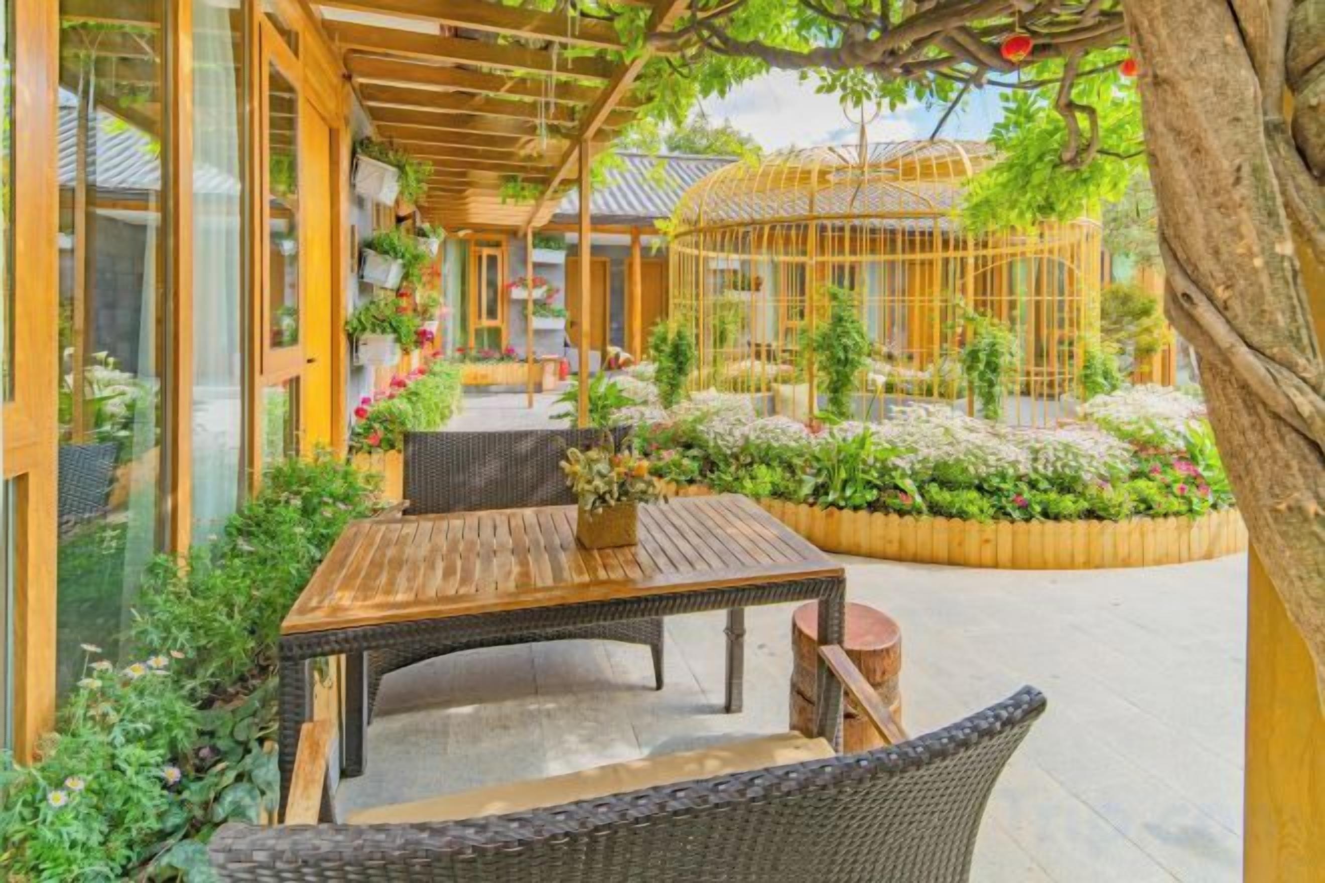 An outdoor seating area with a wooden table and wicker chairs, surrounded by lush plants and a wooden fence.