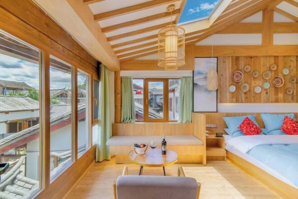 A room with a wooden ceiling, large windows, a bed, a bench, and a table with a bottle and bowl.