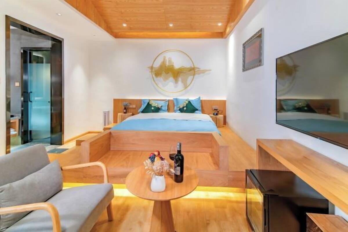 A modern hotel room with a wooden ceiling, a bed with blue bedding, a TV mounted on the wall, and a small table with a bottle and flowers.