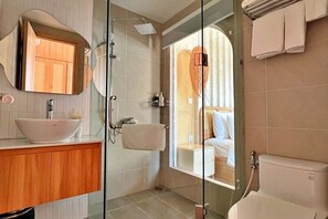 Un baño moderno con cabina de ducha de vidrio, tocador de madera y vista a un dormitorio a través del vidrio.