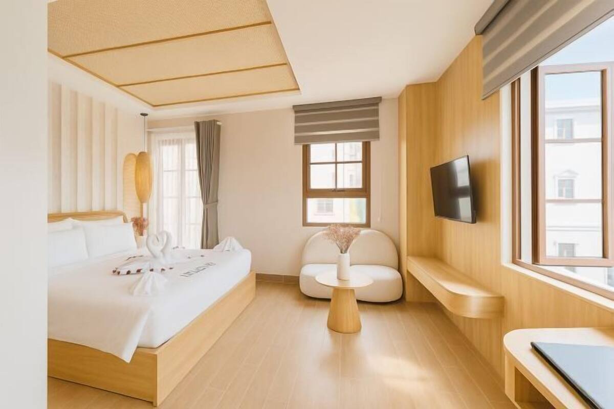 A modern hotel room with a large bed, a seating area, a TV, and a window with a view of buildings.
