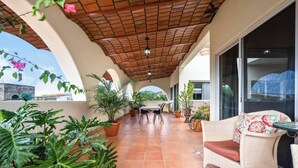 Um alpendre coberto com azulejos de barro, vasos de plantas, uma mesa de jantar e uma cadeira de vime.