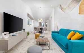 Ein modernes Wohnzimmer mit einem großen Flachbildfernseher, einem türkisfarbenen Sofa mit gelben Kissen, einem Couchtisch und einem Essbereich mit Stühlen und einer Chandelüster.