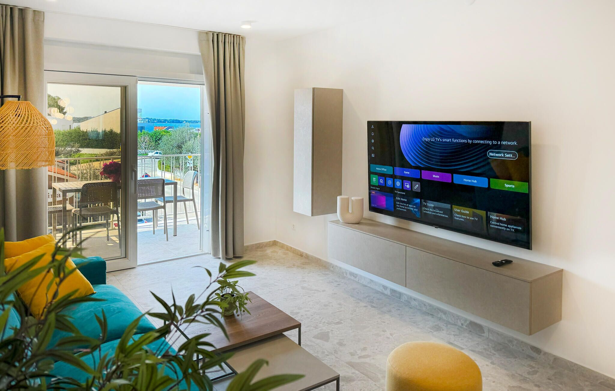 A modern living room with a flat-screen TV mounted on the wall, a sofa, a coffee table, and a balcony with outdoor seating.