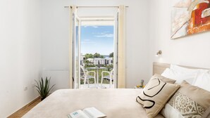 Quarto com cama, um livro e vista para uma varanda com mobiliário de exterior.