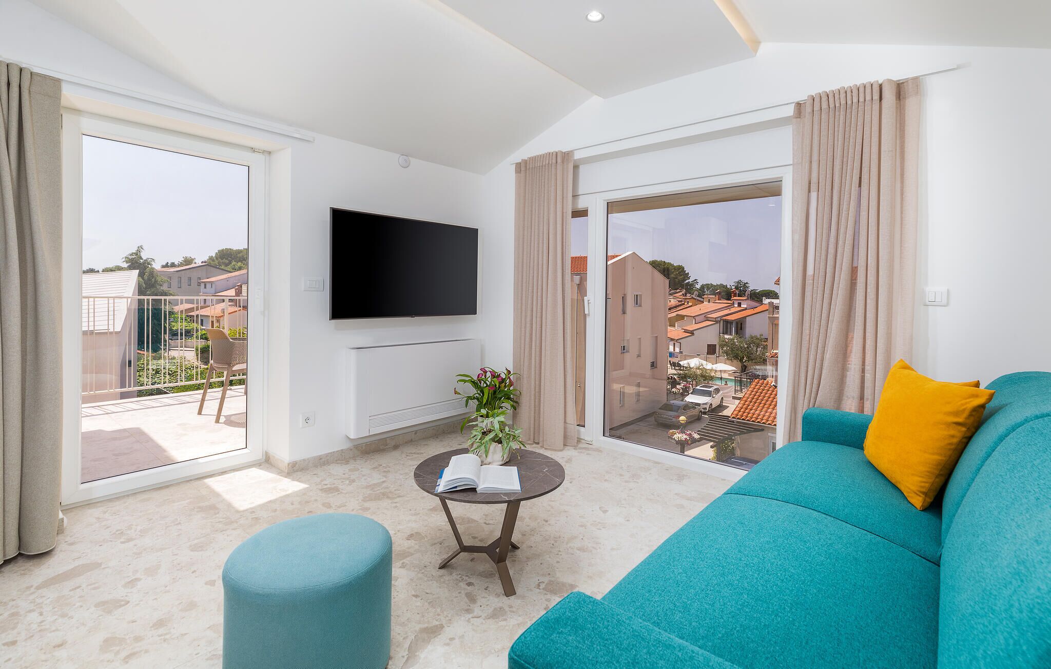 A modern living room with a teal sofa, a flat-screen TV, a small table with a book, and a view of a residential area through large sliding doors.