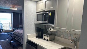 A compact kitchen with a microwave, toaster, and sink. A balcony with a chair and table is visible in the background. - Amazing Beach Location and Ocean View! Unit 220 (Daytona Beach)
