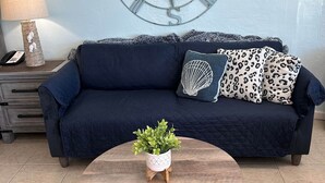 Un soggiorno con un divano blu scuro, cuscini a stampa leopardata, un tavolino rotondo in legno con una pianta e una sveglia a parete.