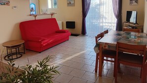 A living room with a red sofa, a dining table with chairs, a TV, and a window with curtains. - T2 48m² Soulac sur mer (Soulac-sur-Mer)