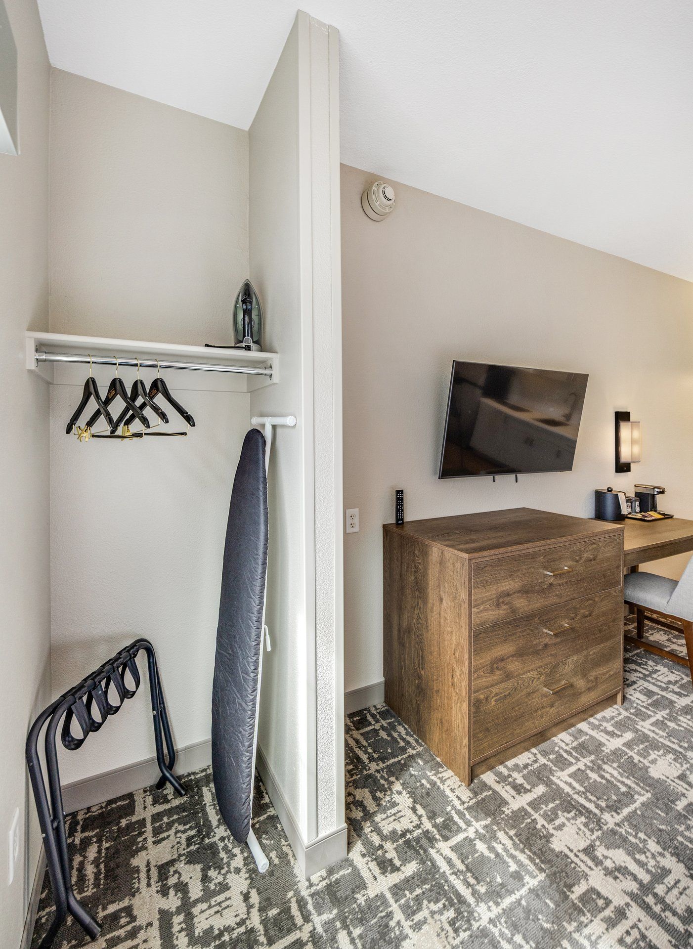 A hotel room with a wooden dresser, a flat-screen TV mounted on the wall, a towel rack with hangers, and a ironing board.