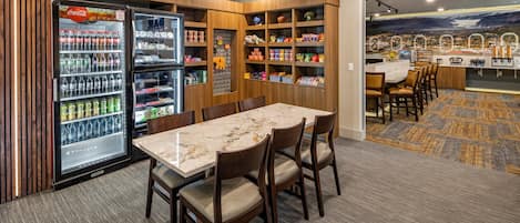 Un comedor de hotel con una mesa de mármol, sillas, un refrigerador lleno de bebidas y estantes con aperitivos.