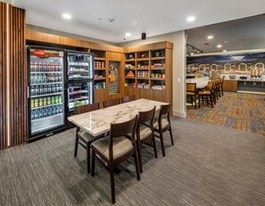 Een restaurantgedeelte in een hotel met een marmeren tafel, stoelen, een koelkast gevuld met drankjes en planken met snacks.