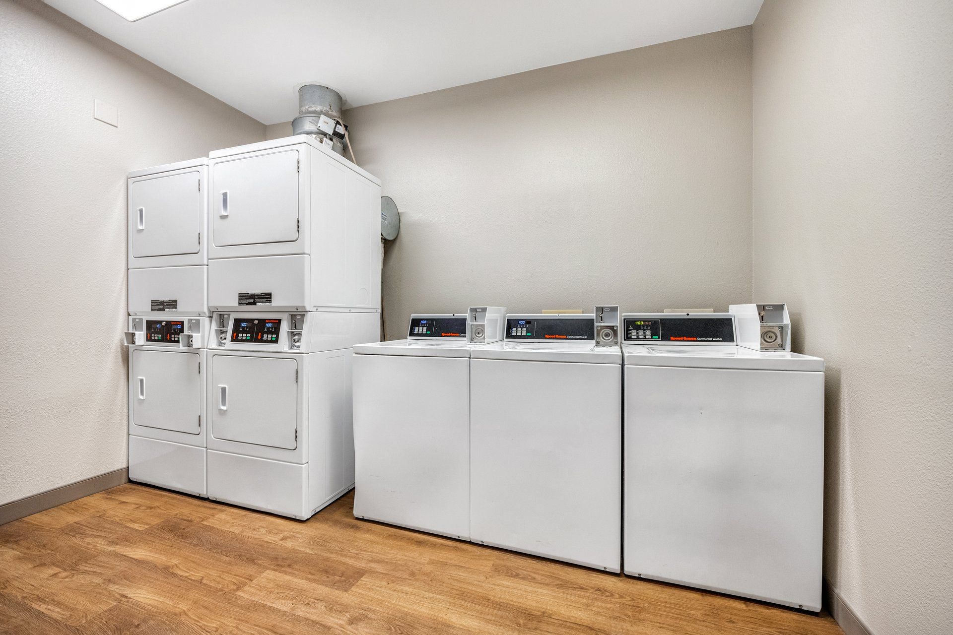 A laundry room with stacked washers and dryers, a vent on the ceiling, and wooden flooring.