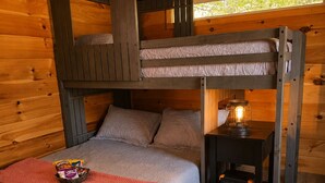 A bunk bed room with a small table and lamp, wooden walls, and a window.