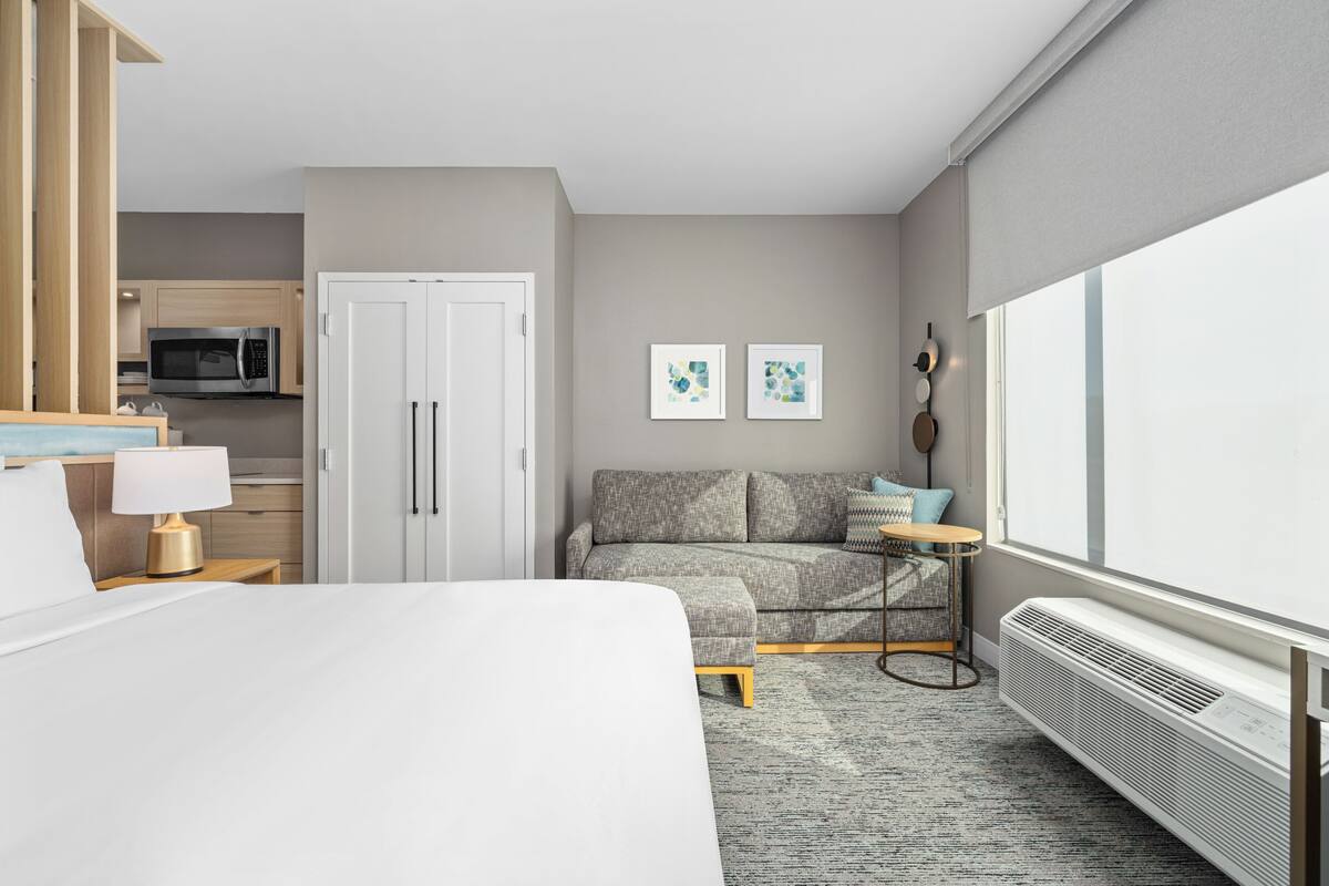 Habitación de hotel con cama, sofá, una mesita, lámpara, microondas, refrigerador y un ventanal grande.