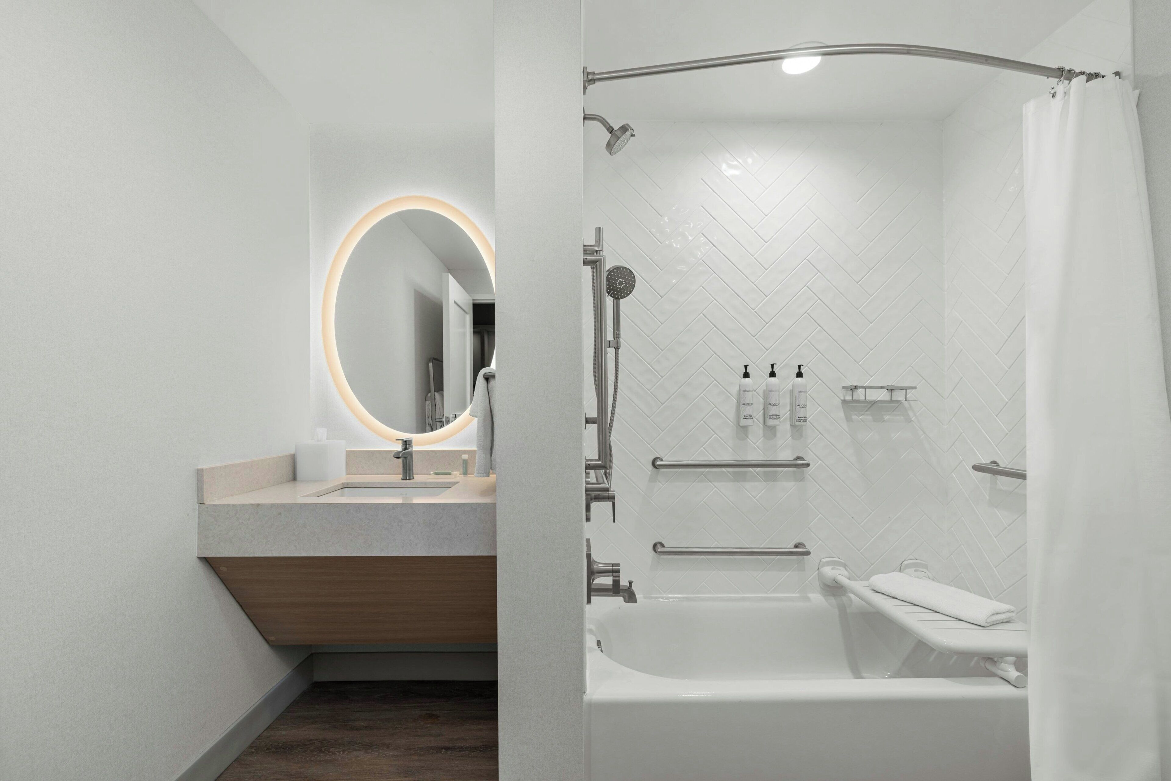 Un baño moderno con tocador, un espejo con luz circular y una ducha con puerta de vidrio curva, además de un patrón de baldosas tipo espiga en las paredes.