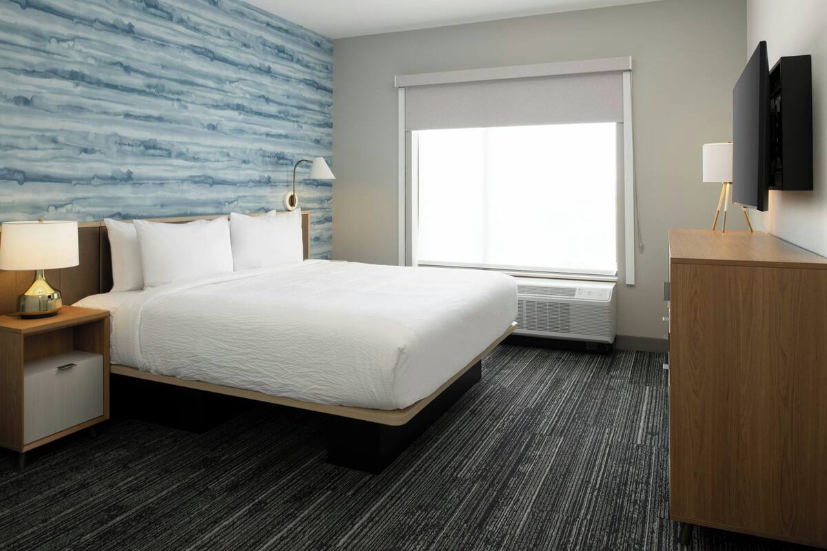 Habitación de hotel con cama, mesita de noche con lámpara, cómoda, ventana grande y televisor montado en la pared.