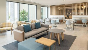 Un hall d’hôtel moderne, doté d’un canapé, de fauteuils, de tables et d’une grande fenêtre donnant sur un paysage verdoyant.