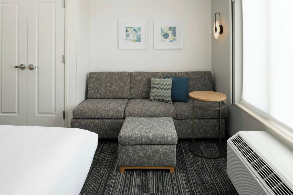 Una habitación de hotel moderna con un sofá gris, una mesita redonda pequeña, un otoman gris y una cama con edredón blanco. Hay dos obras de arte abstractas enmarcadas en la pared sobre el sofá.