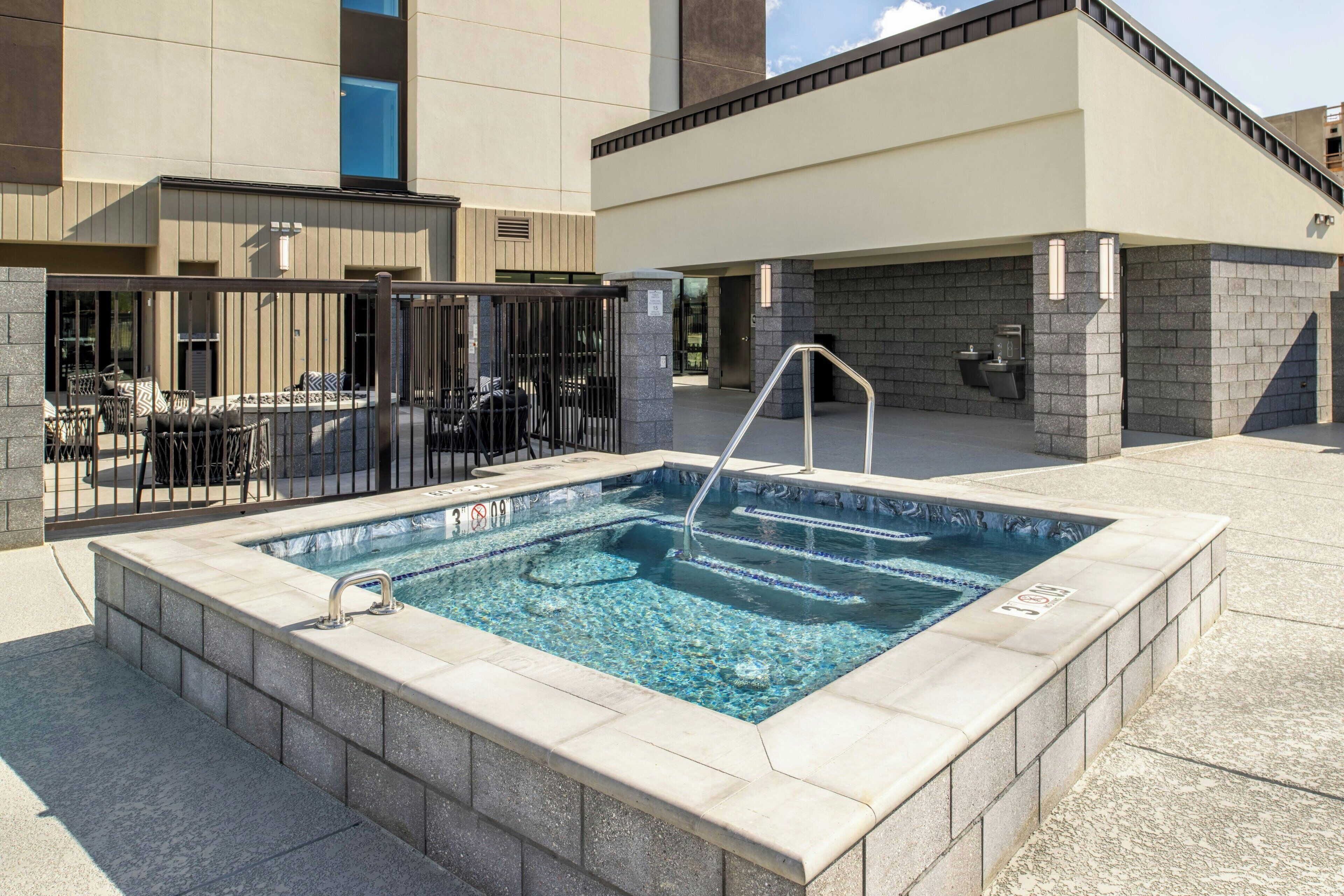 Piscina rectangular al aire libre con una escalera metálica, rodeada de baldosas de piedra, y contigua a un edificio con fachada de baldosas y una zona cubierta con sillas y mesas.