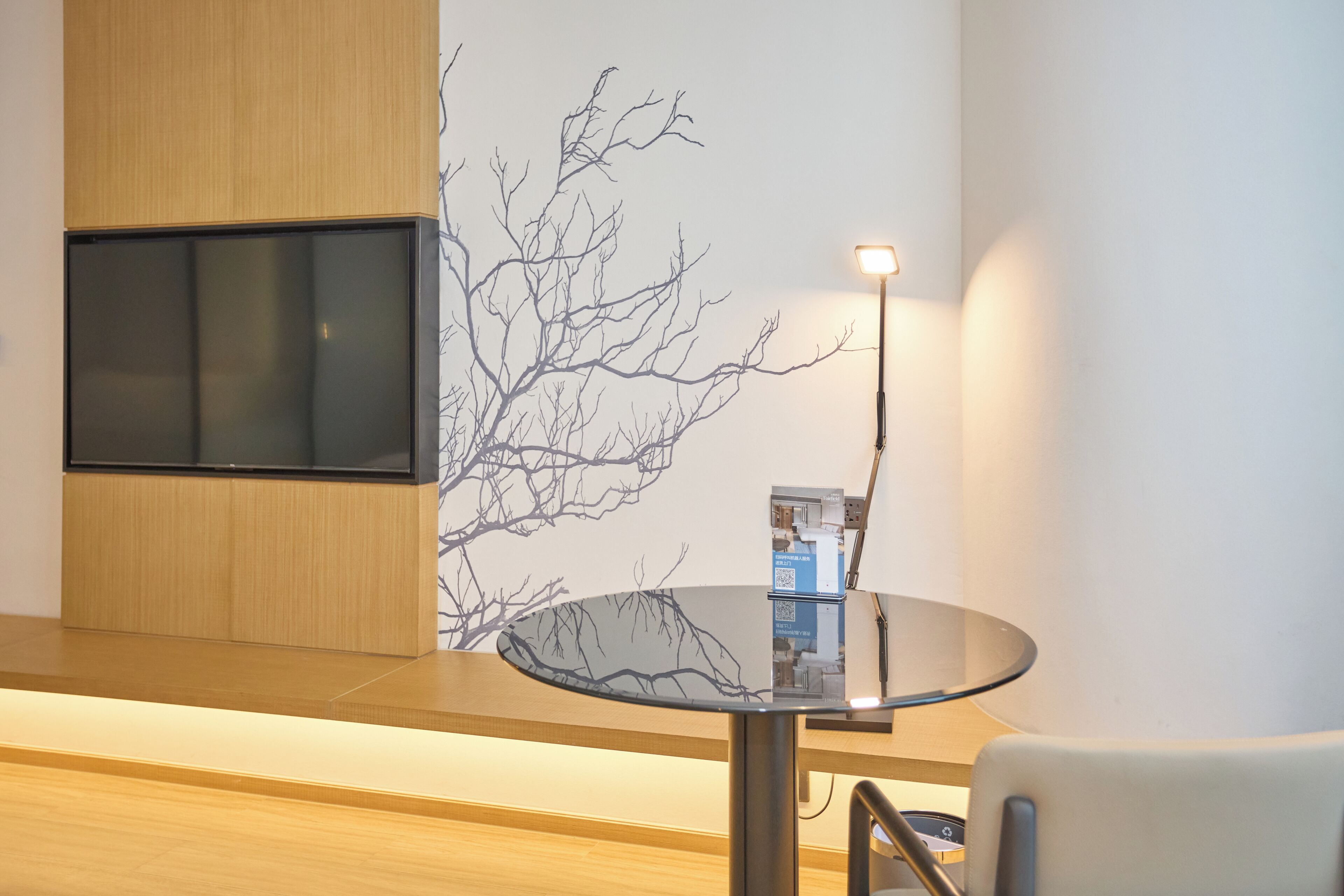 A modern hotel room with a flat-screen TV mounted on a wooden panel, a round glass table with a lamp, and a wall mural of bare tree branches.