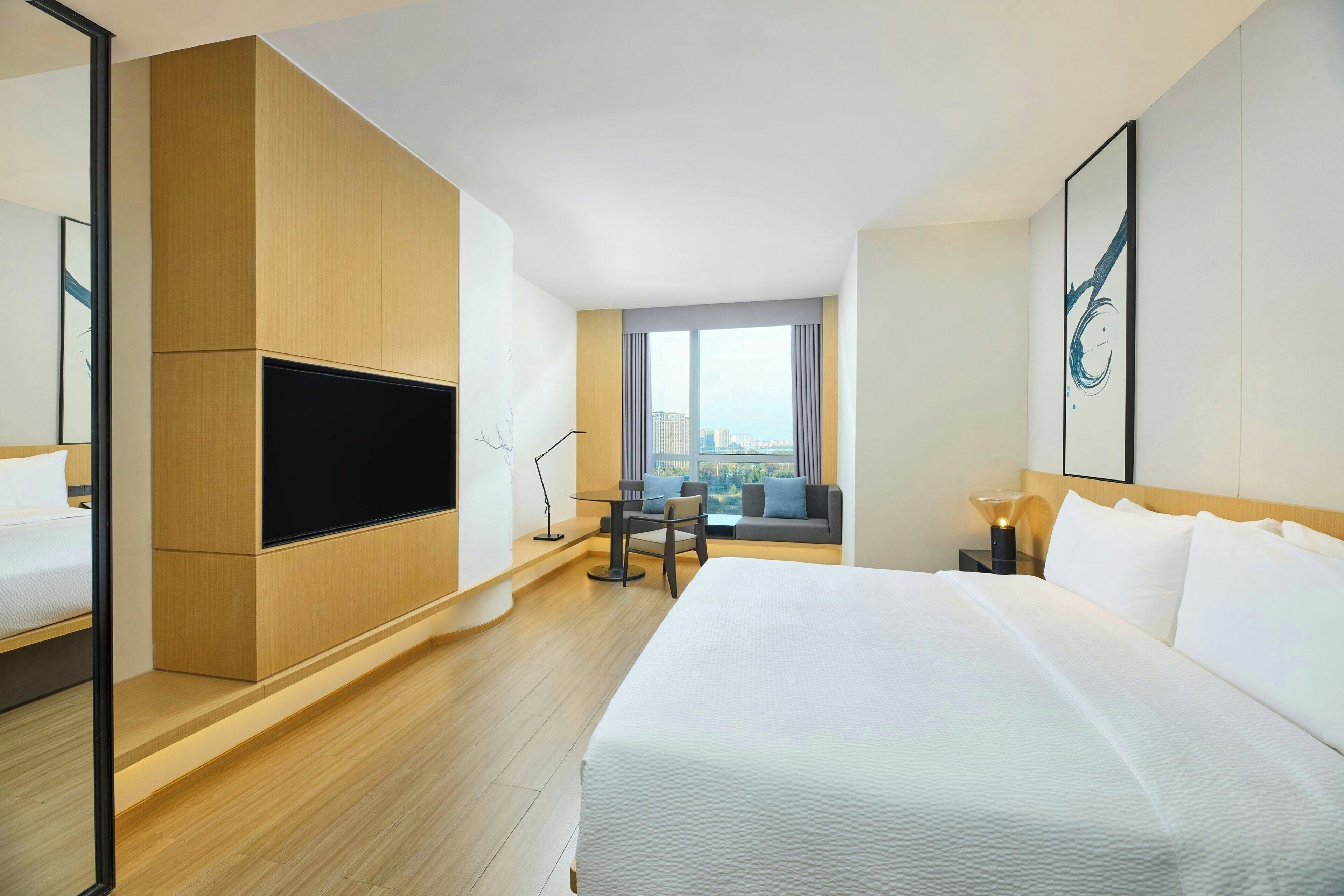 A modern hotel room with a large bed, a flat-screen TV, a seating area with a view, and a cityscape visible through the window.