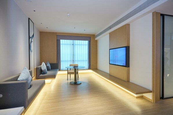Una habitación de hotel moderna con sofá, una pequeña mesa redonda, televisor de pantalla plana en la pared y ventanas amplias.