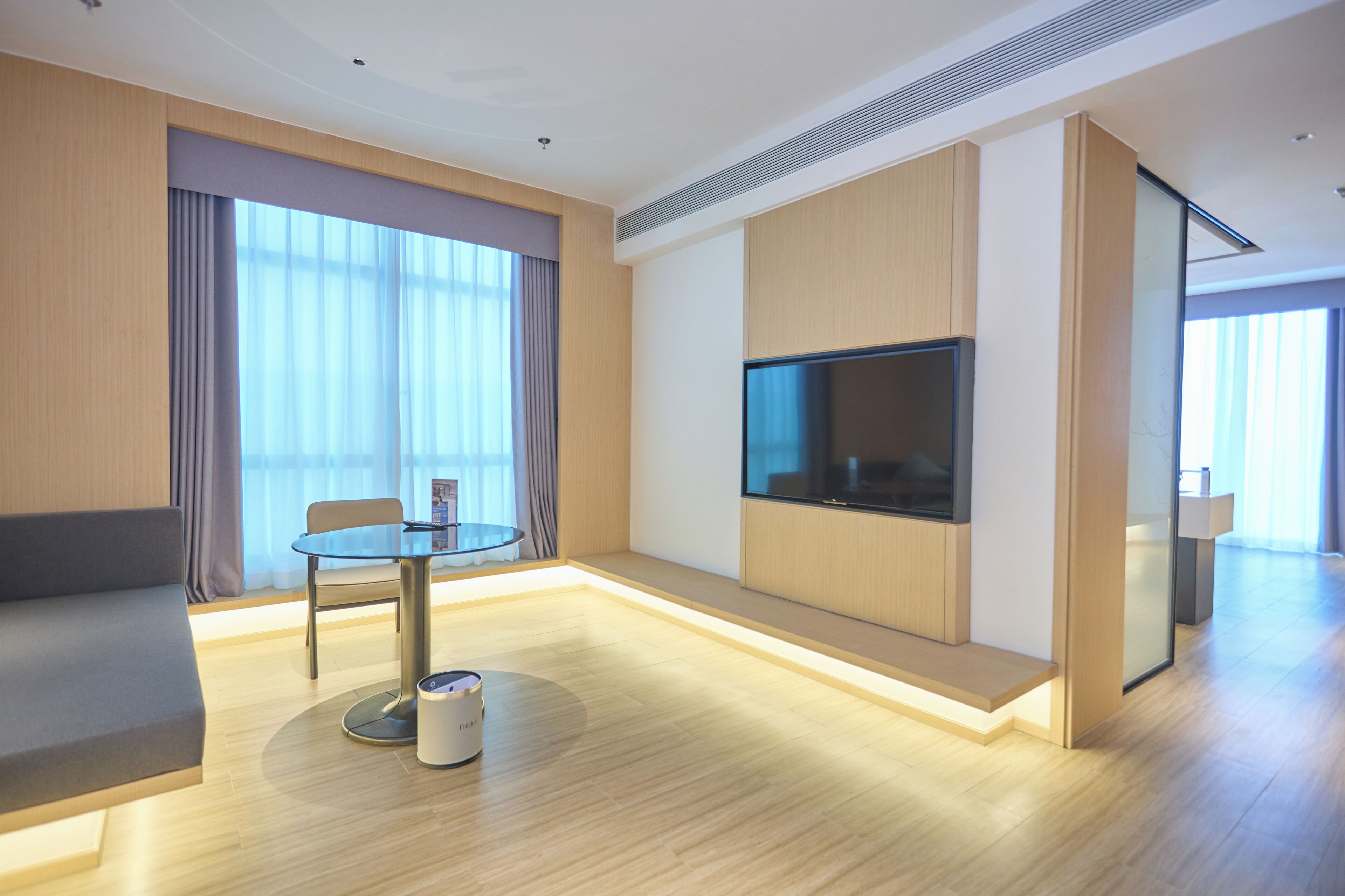 Ett modernt hotellrum med en soffa, ett runt bord med en lampa, en platt-TV och stora fönster med gardiner.